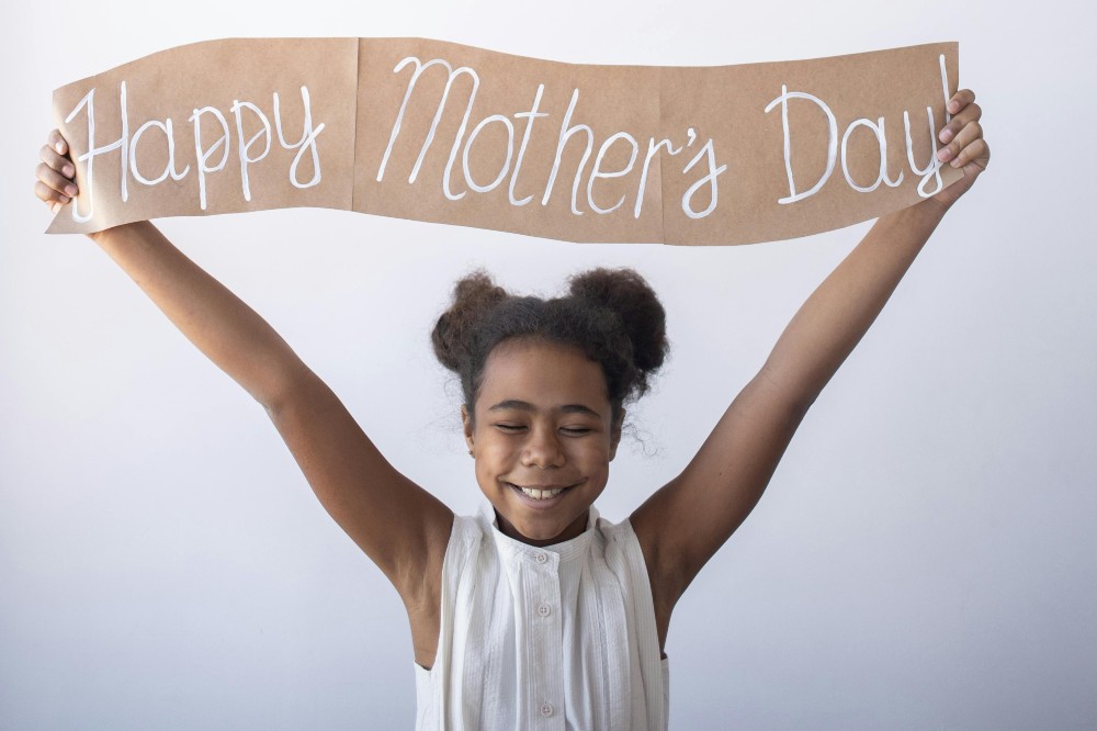 A young girl smiling holding up a paper banner saying Happy Mother's Day!