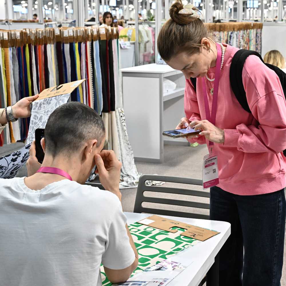 A woman in a pink jumper and black trousers looking down at her phone on a fabric stand at Texhibition