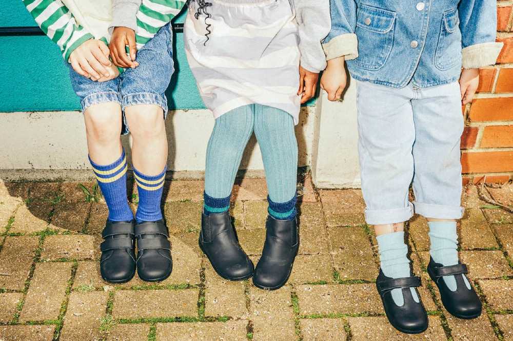 Three children's legs shown stood outside in front of a blue door wearing black school shoes 