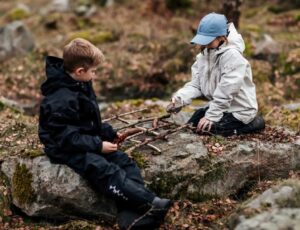 Two children sat outside on a rock playing with sticks wearing outerwear by Isbjorn of Sweden