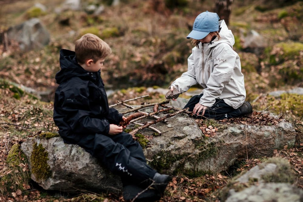 Two children sat outside on a rock playing with sticks wearing outerwear by Isbjorn of Sweden