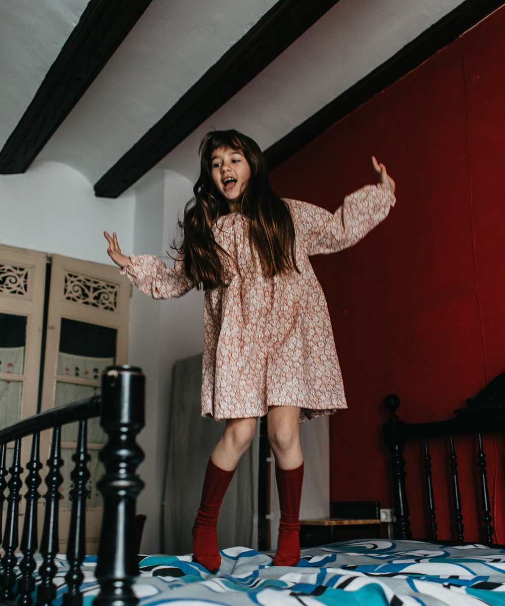 A girl jumping on a bed wearing a dress and maroon knee socks from AliOli Kids Childrenswear Agency 