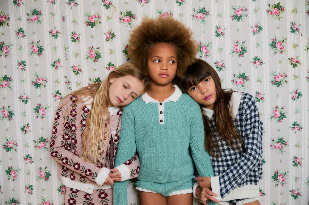 Three girls linking arms stood in front of a wall decorated in floral wallpaper wearing clothes from AliOli Kids Childrenswear Agency