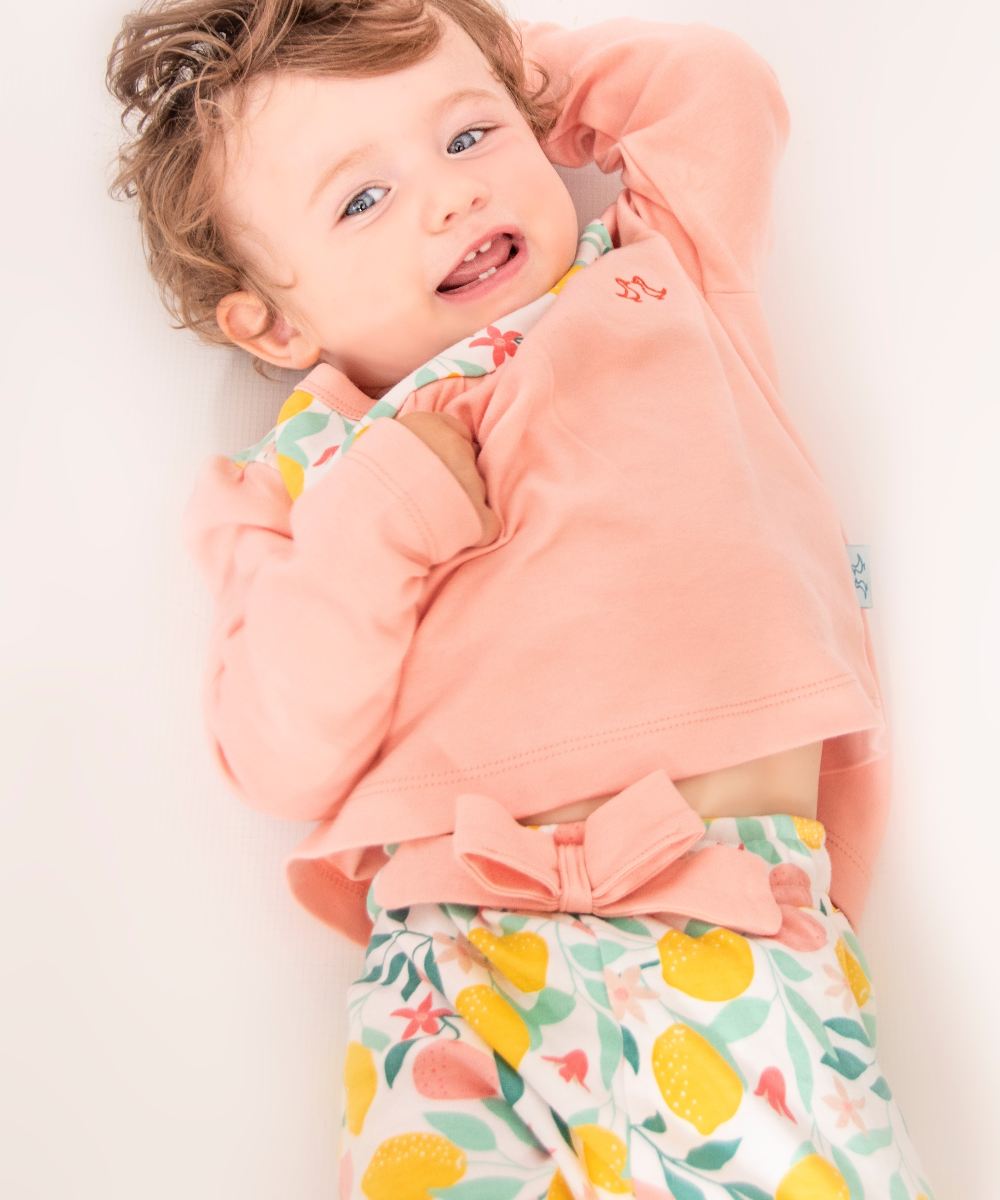 A young child lying on their back smiling wearing a pink top and lemon print trousers by Luca and Rosa London 