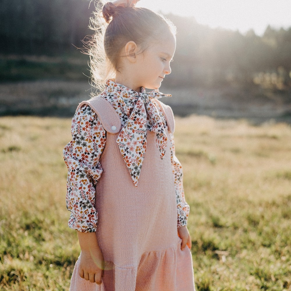 A girl stood outside in a field wearing a pink pinafore dress and floral print shirt with a pussy bow collar 