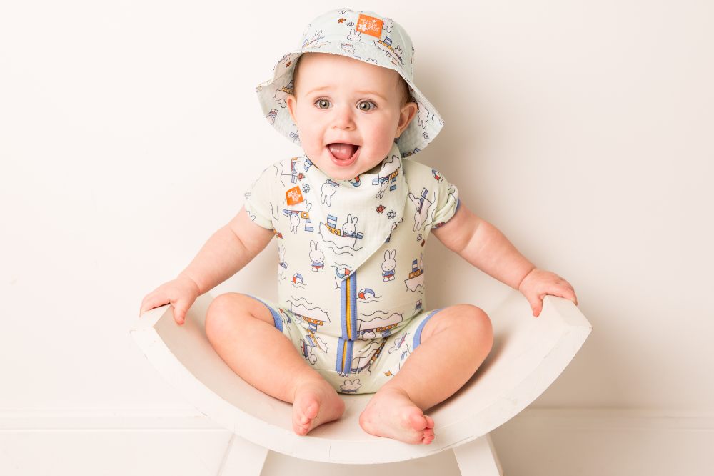 A baby sat in a white chair smiling wearing a swimsuit and hat by Blade & Rose