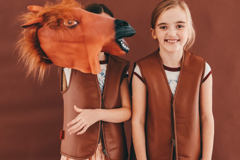 Two girls wearing leather waistcoats with one holding a horses head mask