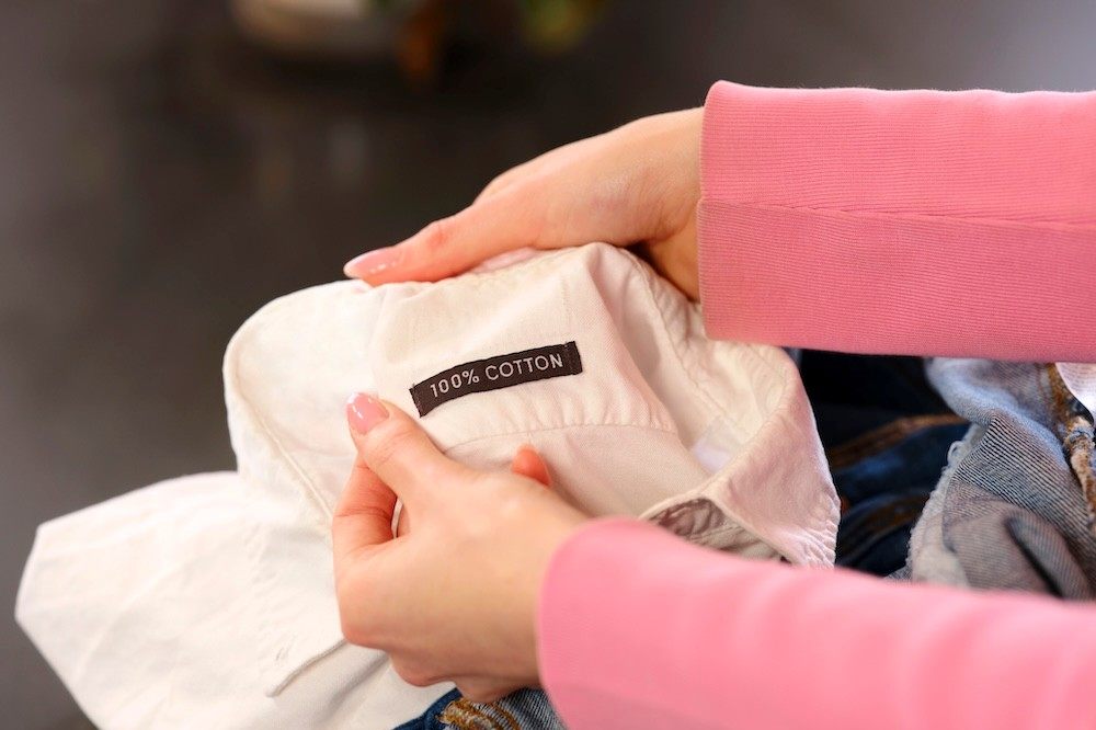 A woman's arms in a pink jumper holding up the neck of a white shirt with a 100% cotton label in it