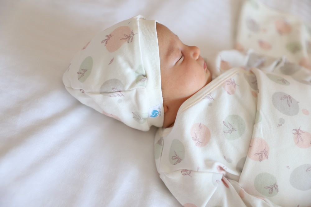A baby lying down asleep on a white sheet wearing a hat and matching babygro
