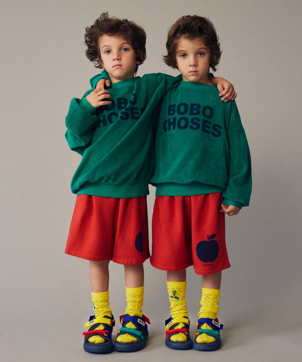 Two identical children wearing matching green jumpers, red shorts, yellow socks and sandals 