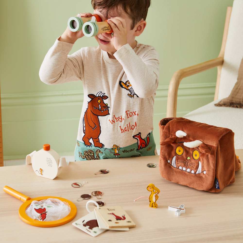 A young boy holding a pair of wooden toy binoculars to his eyes alongside a selection of toys from The Gruffalo by JoJo range 