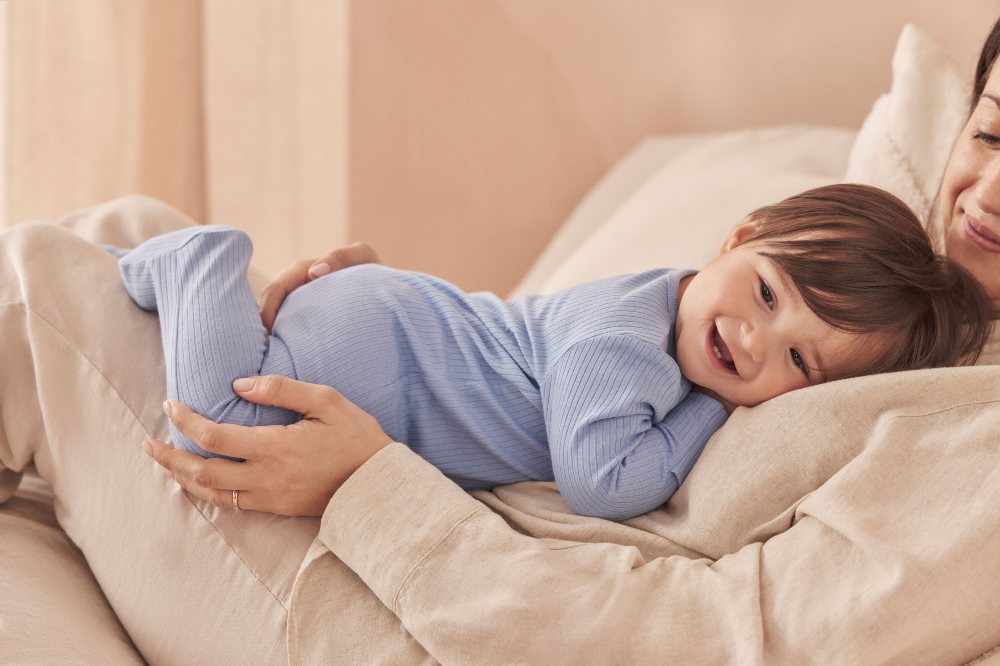 A woman lying down with a young child on her chest wearing a blue babygro by MORI