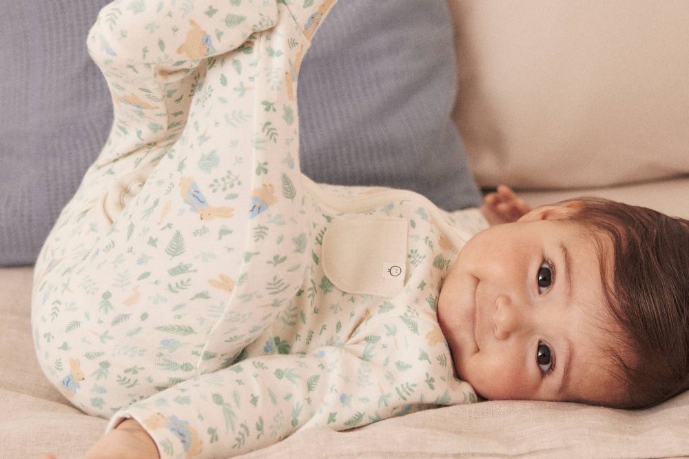 A baby lying with its legs in the ait wearing a babygro by MORI with a Peter Rabbit print
