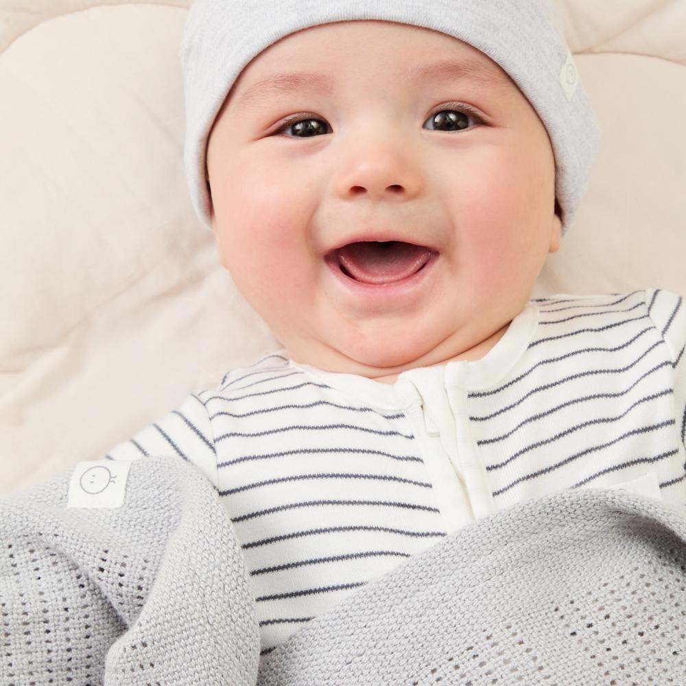 A smiling baby lying down under a blanket wearing a hat, blue and white striped babygro 