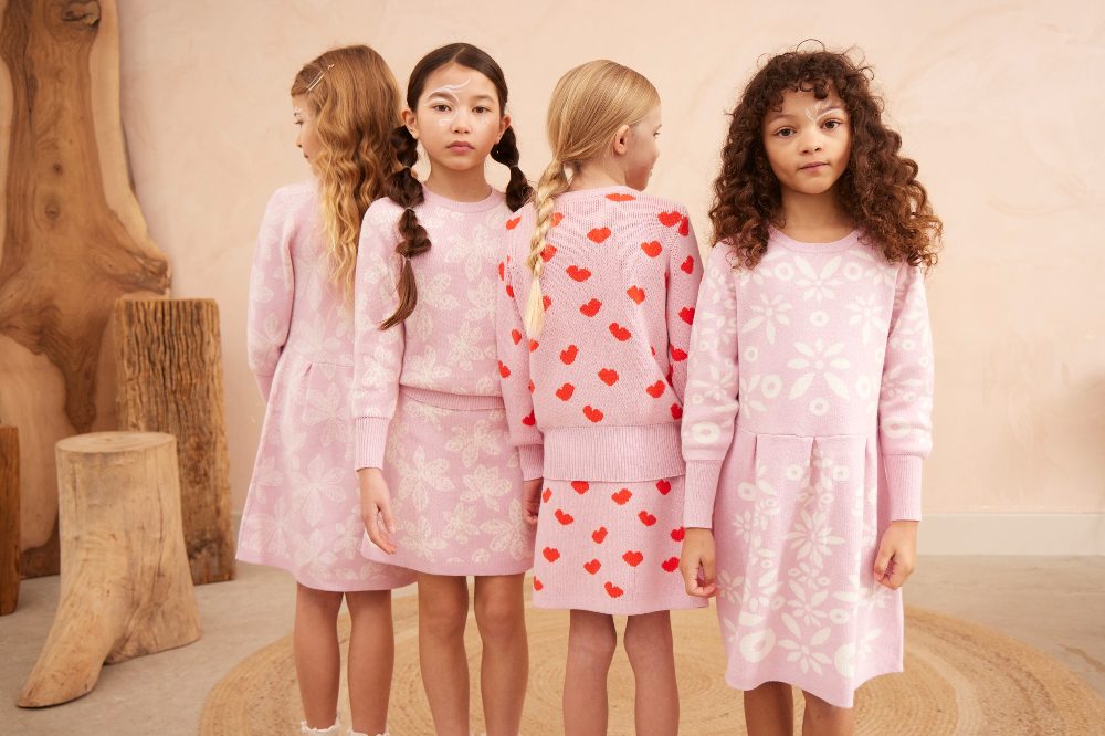 Four girls stood in a row in a room wearing pink knitted outfits by Mika & Milo