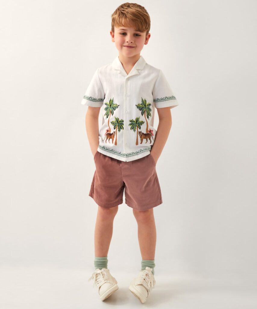 A boy stood in shorts and a short sleeve shirt with palm trees and tiger embroidery by Monsoon Kids 