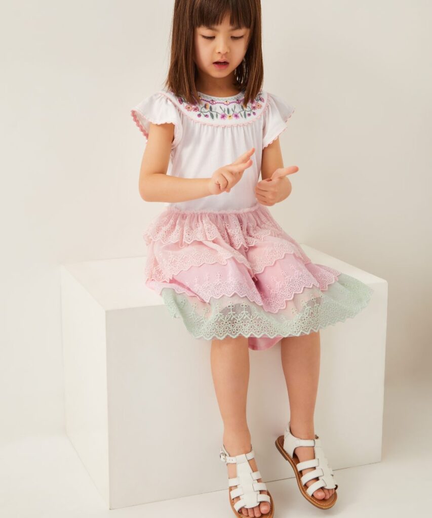 A girl in a white top and pink skirt by Monsoon Kids sitting on a white cube 