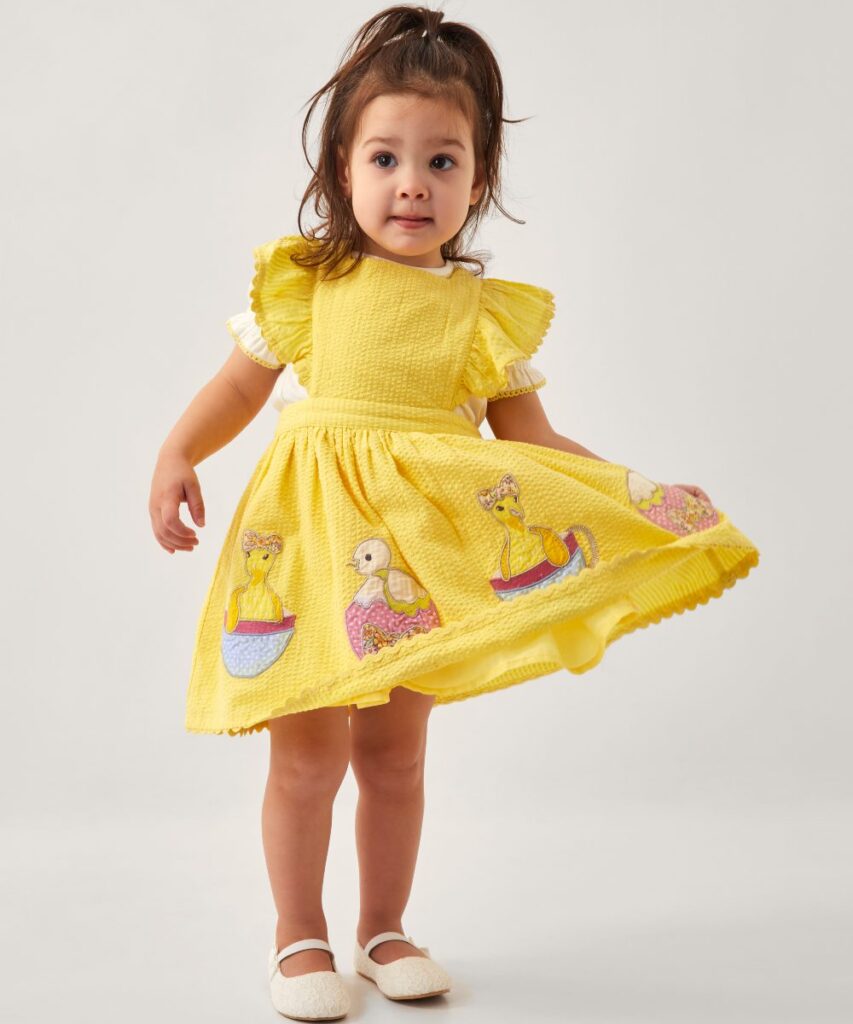 A young girl holding up the corner of her yellow dress with Easter eggs and chicks embroidery on it 