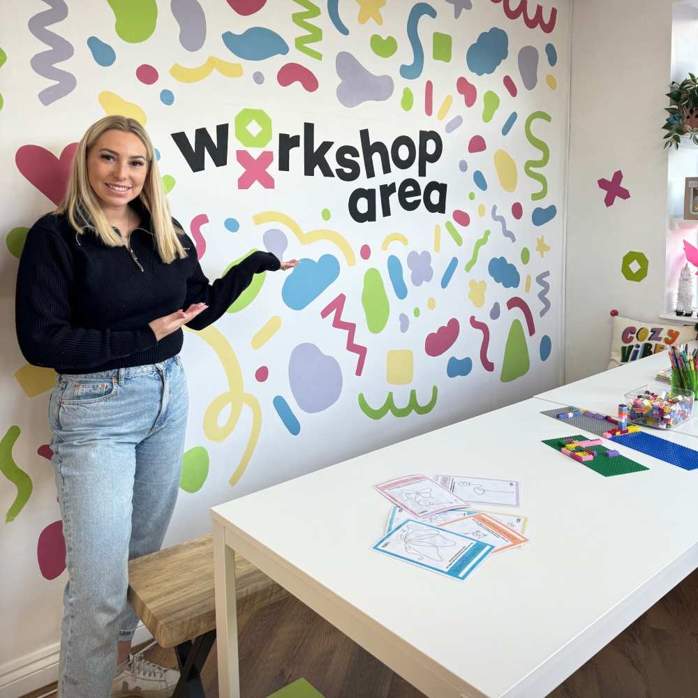 A woman stood beside a wall with workshop area written on it and a craft table in front of her 