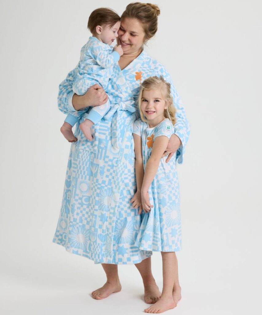A woman in a bathrobe holding a baby and hugging a young girl with them all wearing matching blue and white pieces from the PO.P x Moomins collaboration 