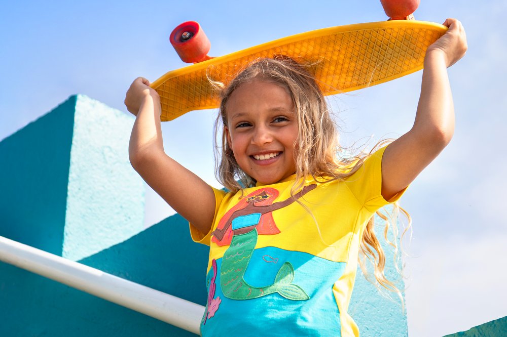 A girl stood outside in the sunshine against a blue wall wearing a T-shirt by Frugi with a mermaid motif and holding a yellow skateboard above her head