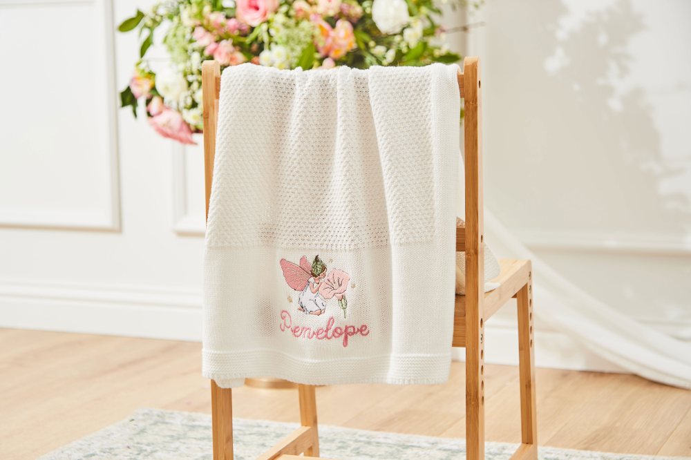 A baby blanket hung on the back of a chair with Penelope and a Flower Fairies motif on it 