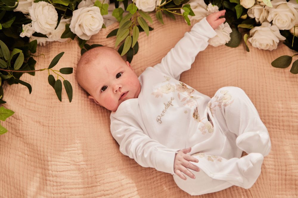 A baby lying beside flowers wearing a babygro with a Flower Fairies motif 