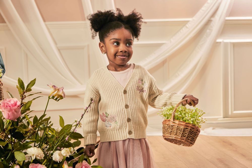 A young girl stood beside a flower display and holding a basket of flowers wearing a knitted cardigan with Flower Fairies motifs