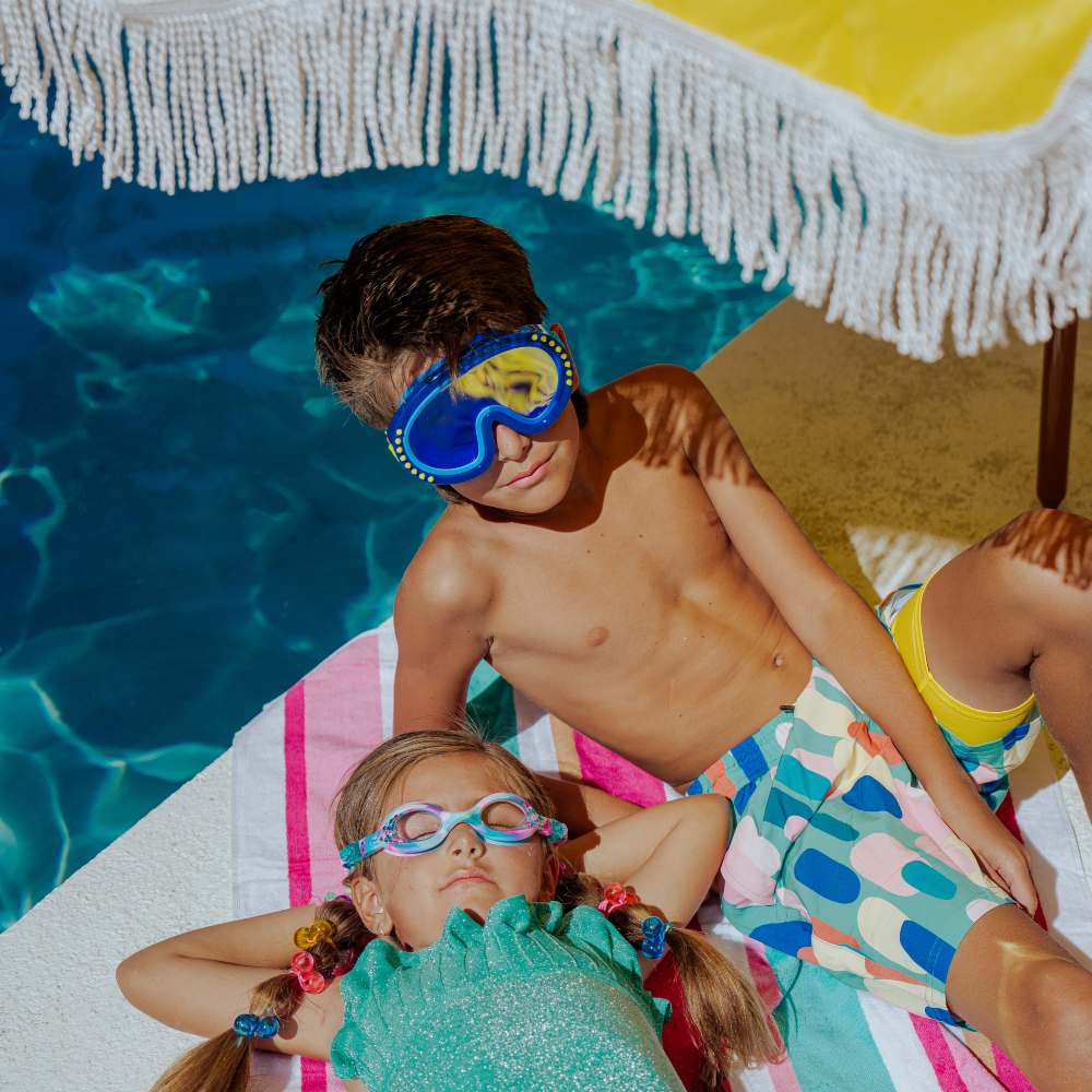 A boy and girl in swim goggles beside a pool underneath a yellow sun shade