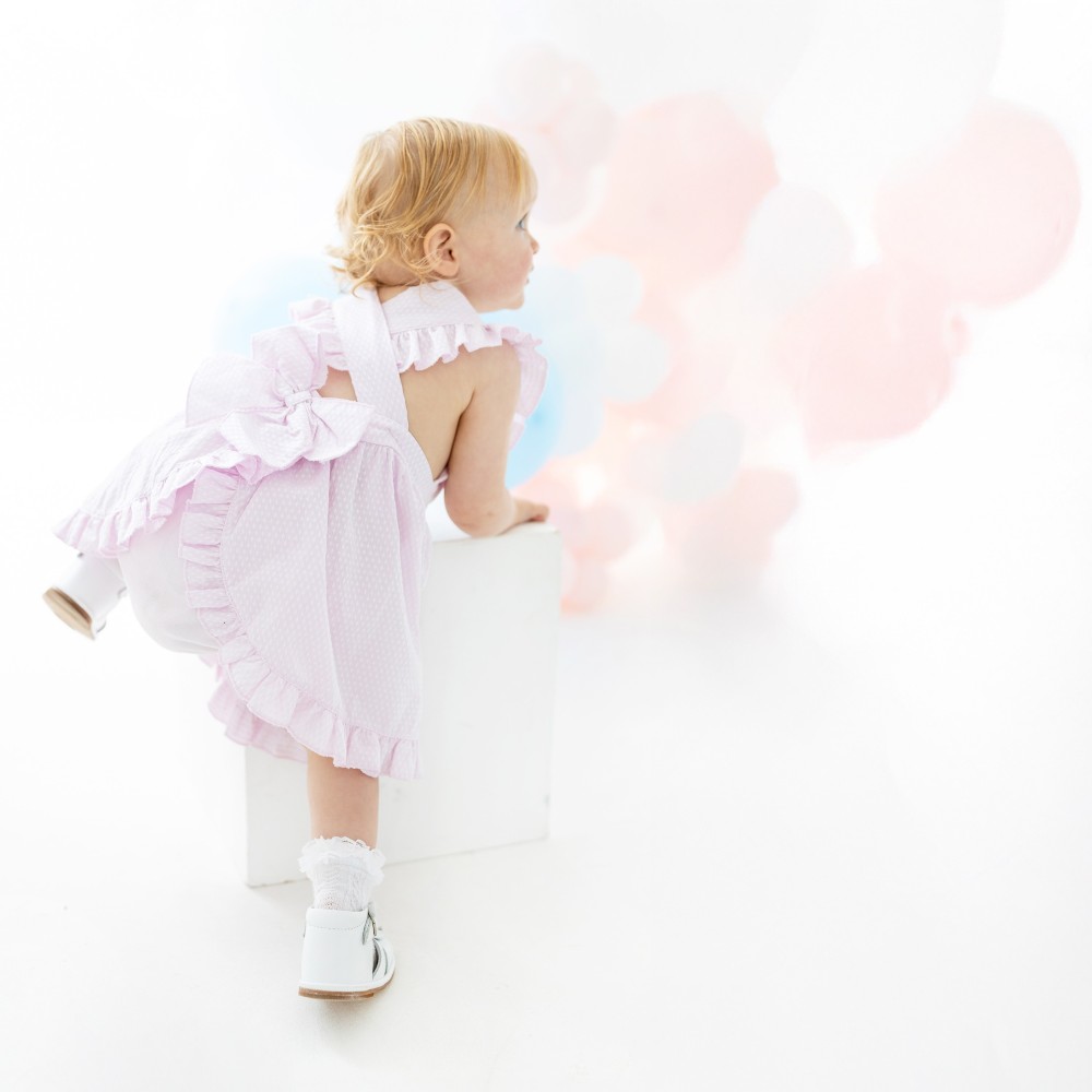 A young girl in a pink, frilly cross back dress climbing onto a chair beside a bunch of balloons