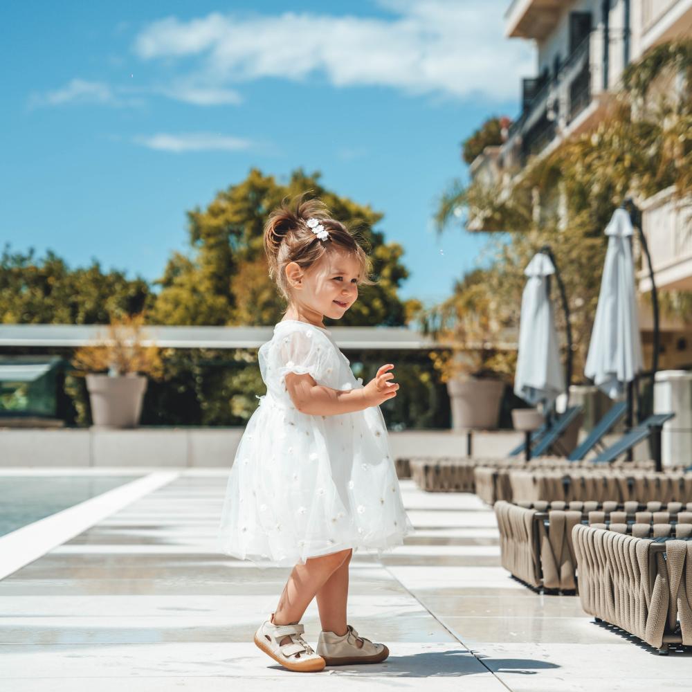 A young girl in a white dress and Froddo shoes stood outside by a swimming pool