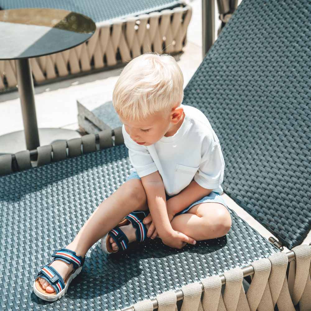 A young boy sat outside on a black sun lounger wearing shorts, a T-shirt and Froddo sandals 