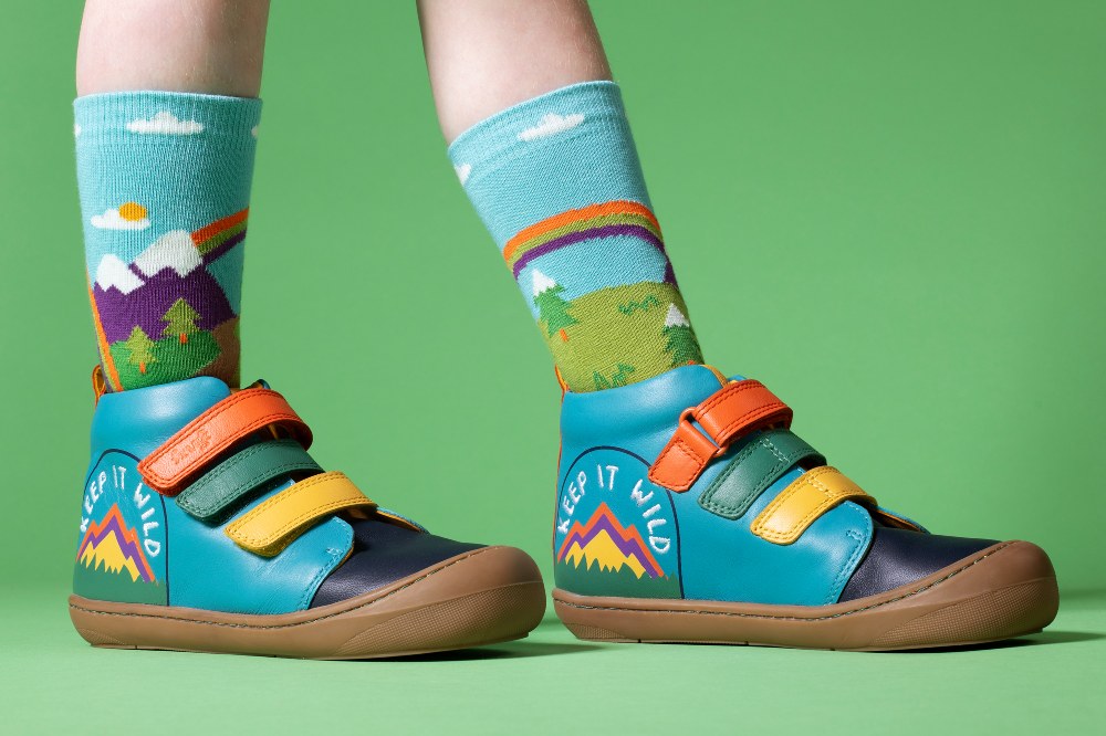 A child's feet wearing socks showing a mountain scene and blue boots with colourful straps by Frugi 
