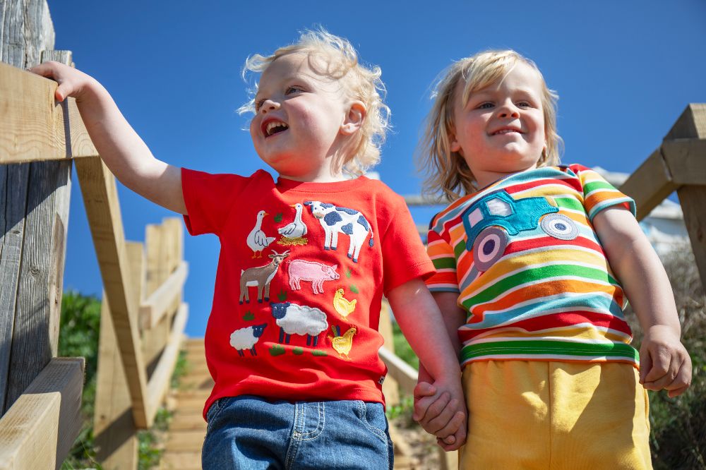 Two young children stood outside in the sunshine holding hands wearing colourful outfits by Frugi 