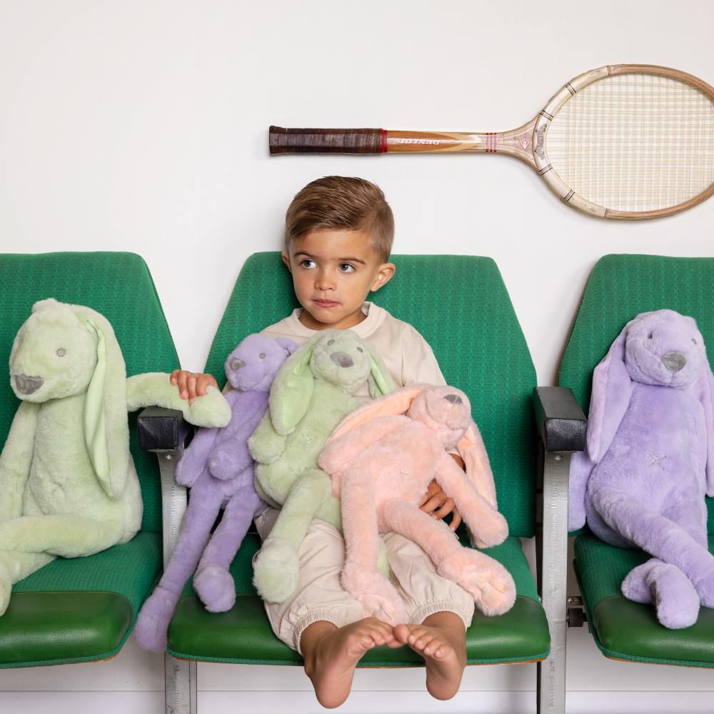 A young boy sat in a green chair holding large rabbit soft toys with a tennis racket above his head 