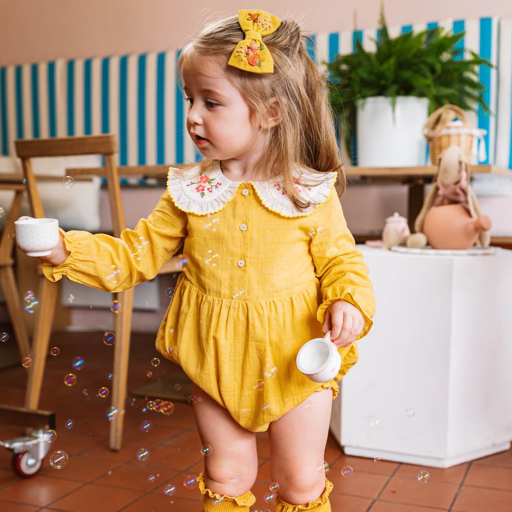 A young girl in a yellow romper with a white collar holding tea cups in a cafe 