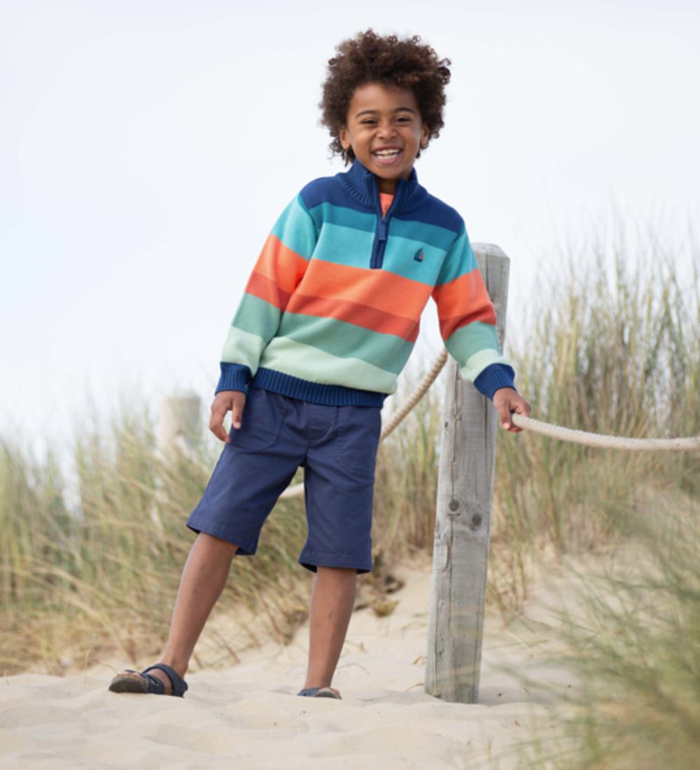 A young boy in a striped jumper and shorts stood on a beach 