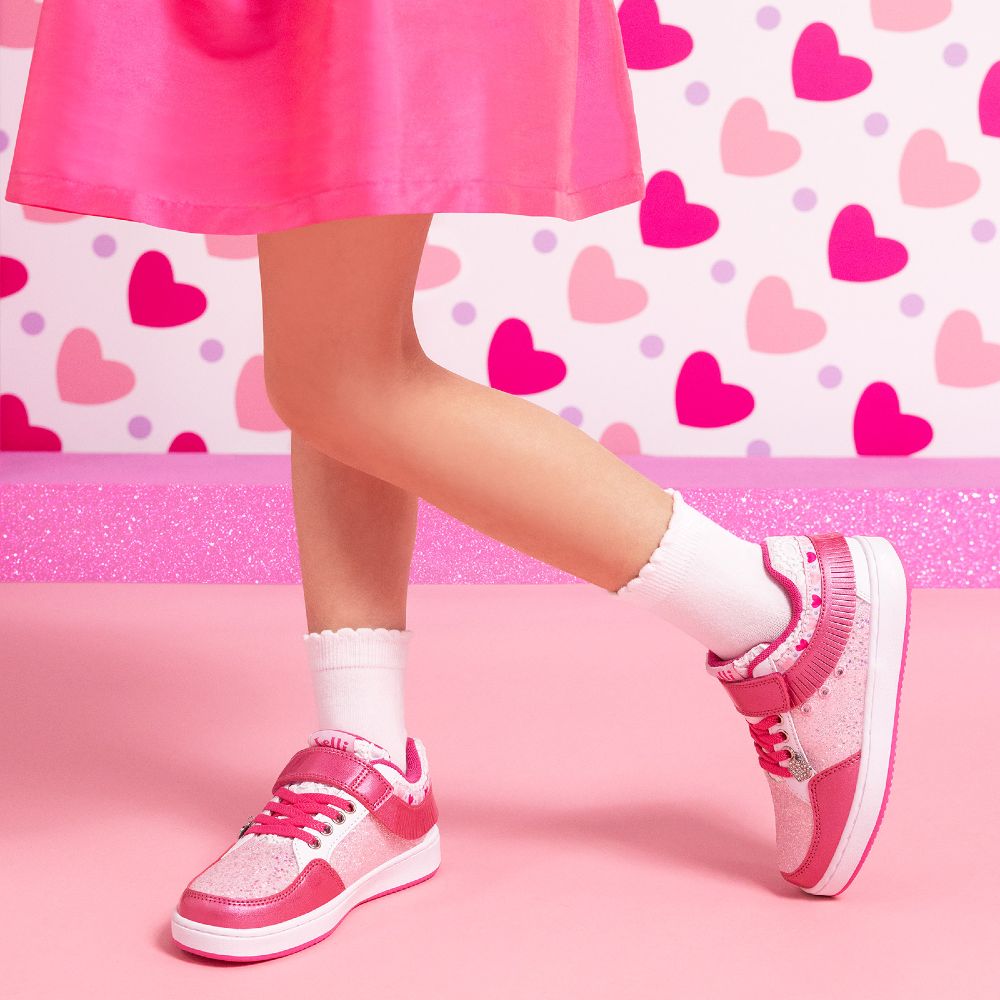 A girl's legs shown wearing a pink skirt and pink Lelli Kelly trainers against a pink heart print wall