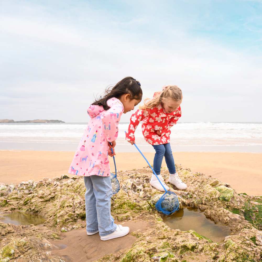 Two children rock pooling on a beach wearing bright patterned raincoats 