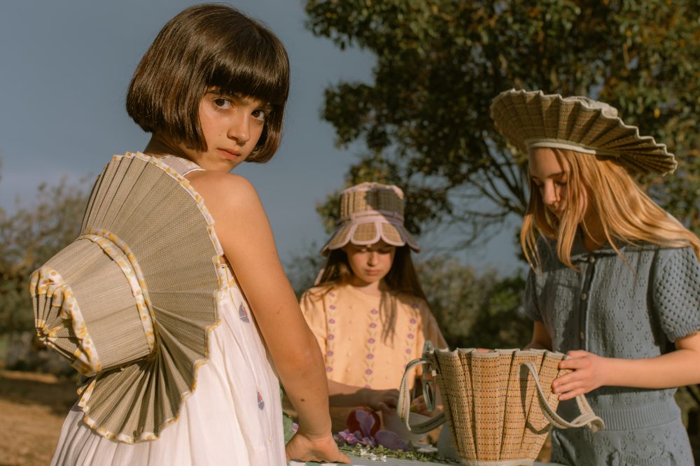 Three girls stood outside at a table wearing hats by Lorna Murray