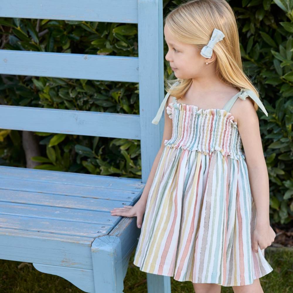 A young girl in a striped summer dress stood outside next to a blue wooden chair 