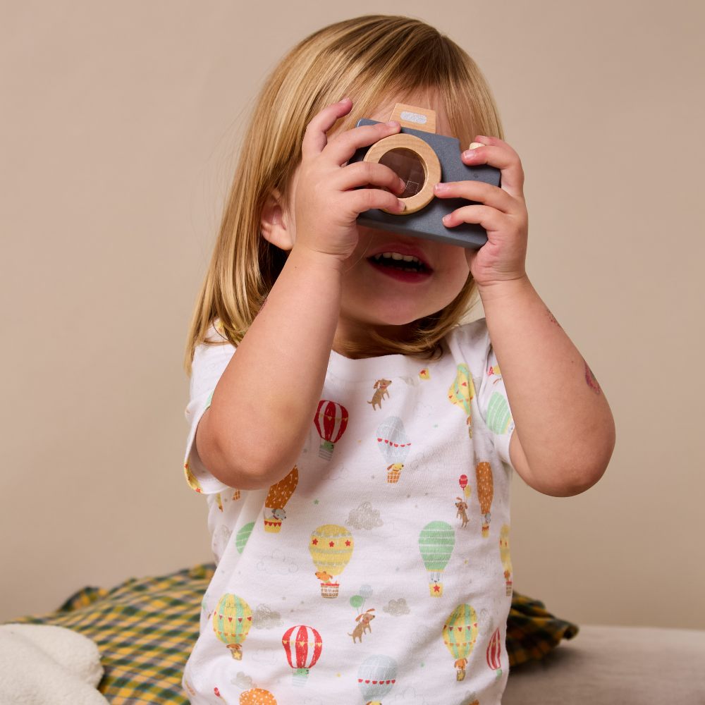 A young girl wearing a T-short with hot air balloons on holding up a wooden toy camera to her face 