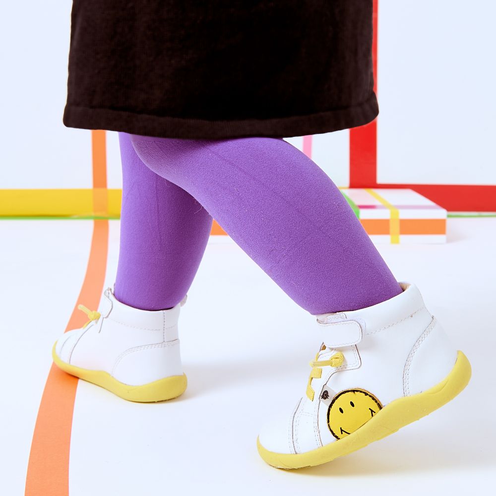 A child's legs shown wearing a black skirt, purple tights and a pair of white trainers with a yellow smiley face motif
