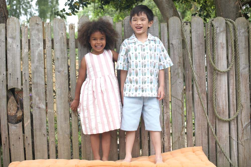 Two young children outside stood on a orange cushion in front of a wooden fence