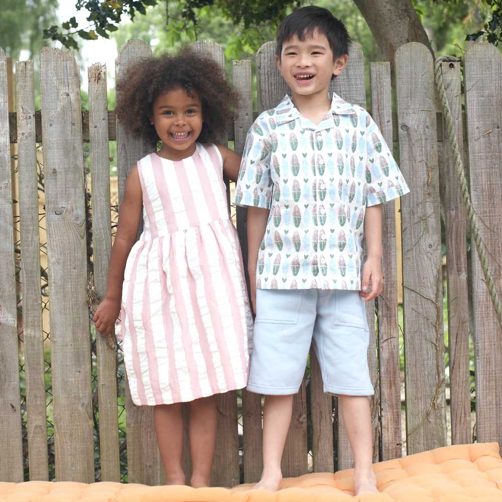Two children in summer outfits stood outside on an orange cushion in front of a fence 