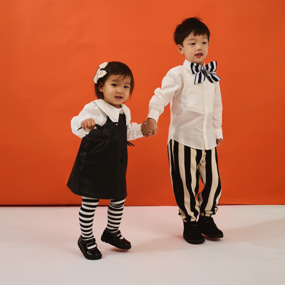 Two children holding hands against an orange background wearing black and white outfits and shoes by Prenta 