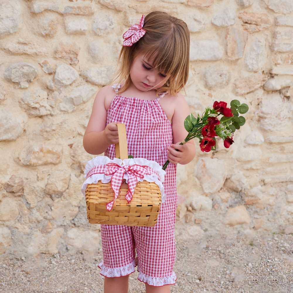 A young girl outside wearing a red gingham outfit holding a basket and a bunch of red flowers