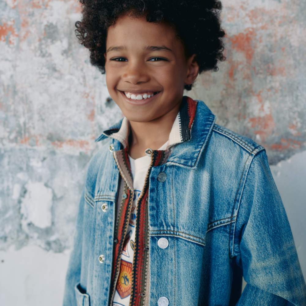 A boy stood against a wall smiling wearing a denim jacket 