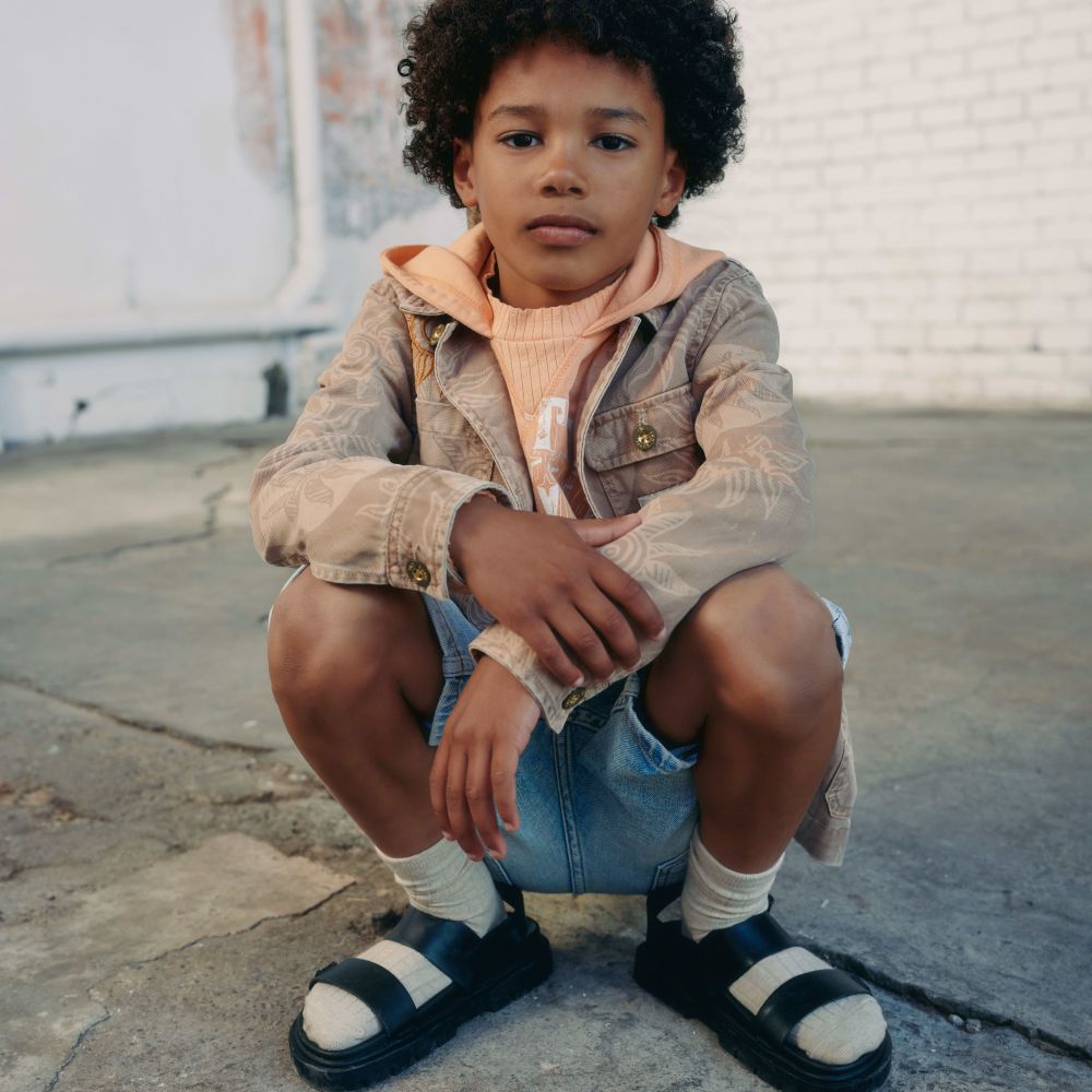A boy crouched down outside wearing a T-shirt, jacket, shorts and black sandals with white socks 