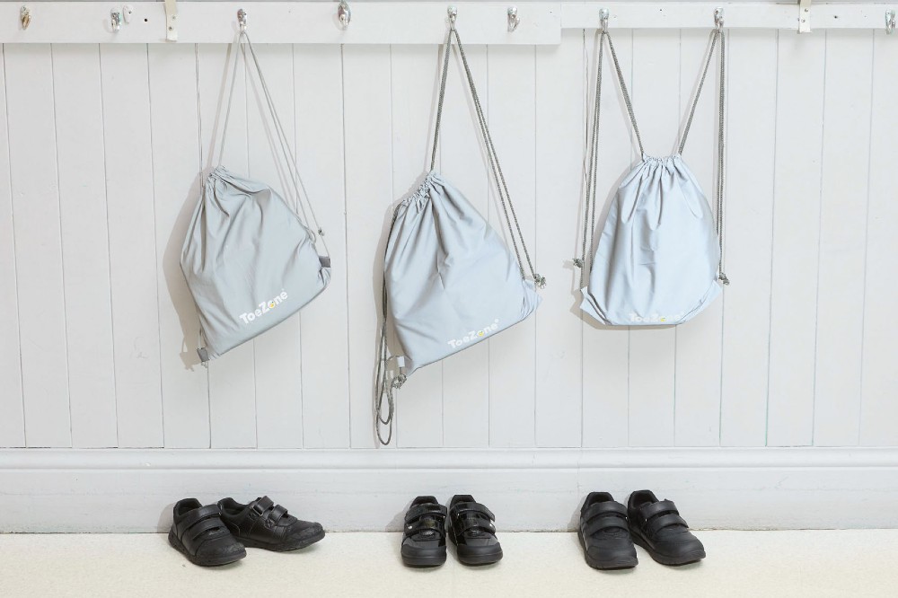 Three pairs of children's black ToeZone school shoes beneath blue bags hung on coat hooks 